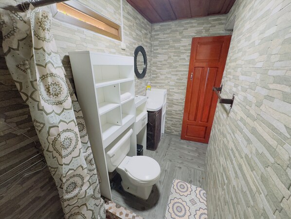 A bathroom with a white toilet, a white shelf unit, a shower curtain with a geometric pattern, a wooden door, and a stone-patterned wall. - Hotel Hanna Suites (Panajachel)