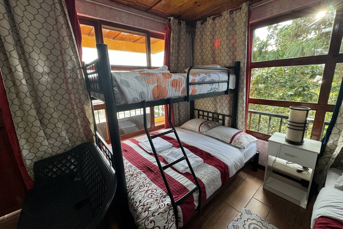 A bunk bed room with a wooden ceiling, patterned curtains, and a window with a view of greenery.
