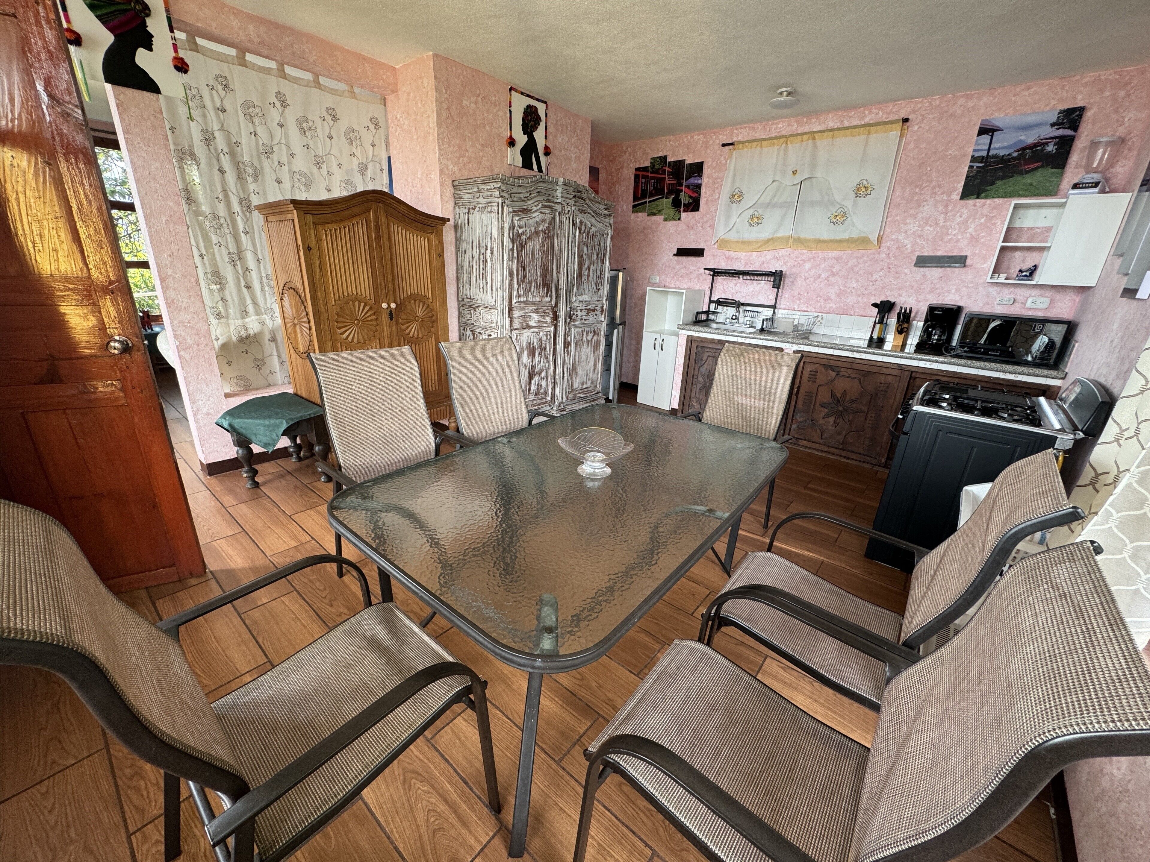 Een eethoek met een glazen tafel en metalen stoelen, een houten kast en een keuken met een fornuis en spoelbak.