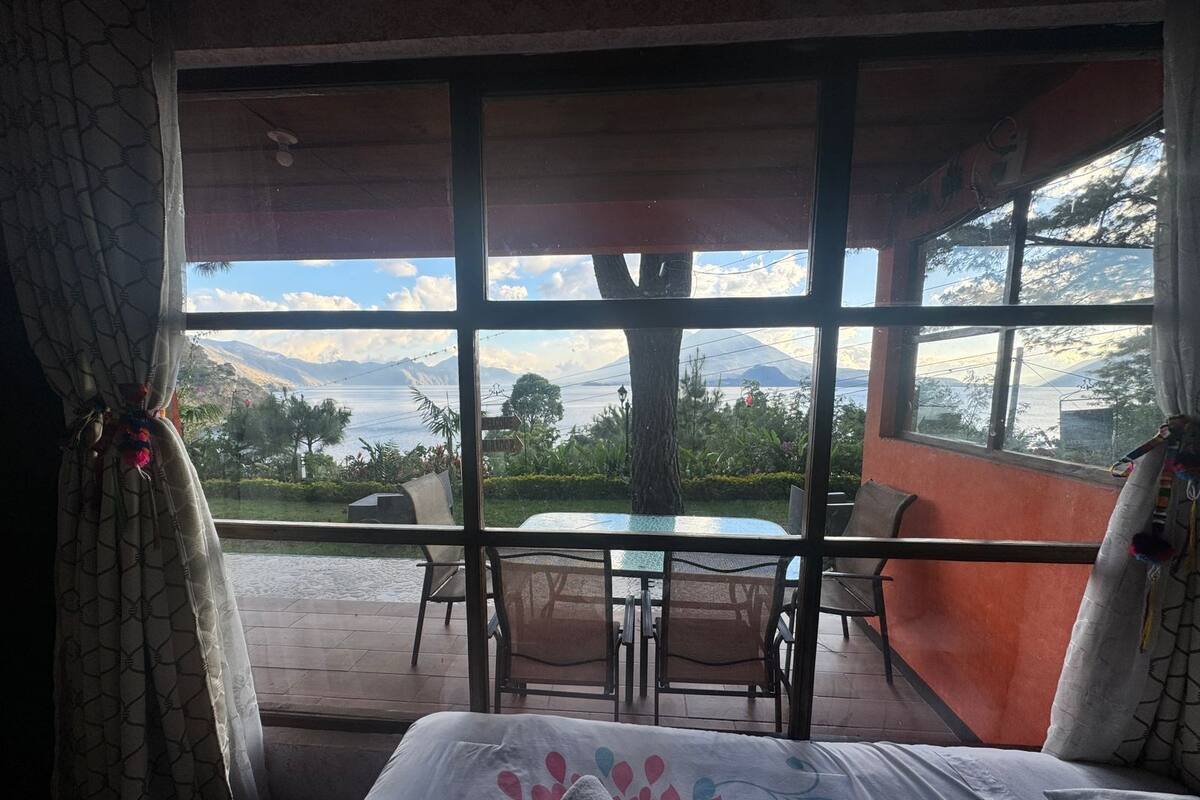 A room with a bed, a table, and chairs, a large window with a view of mountains and greenery.