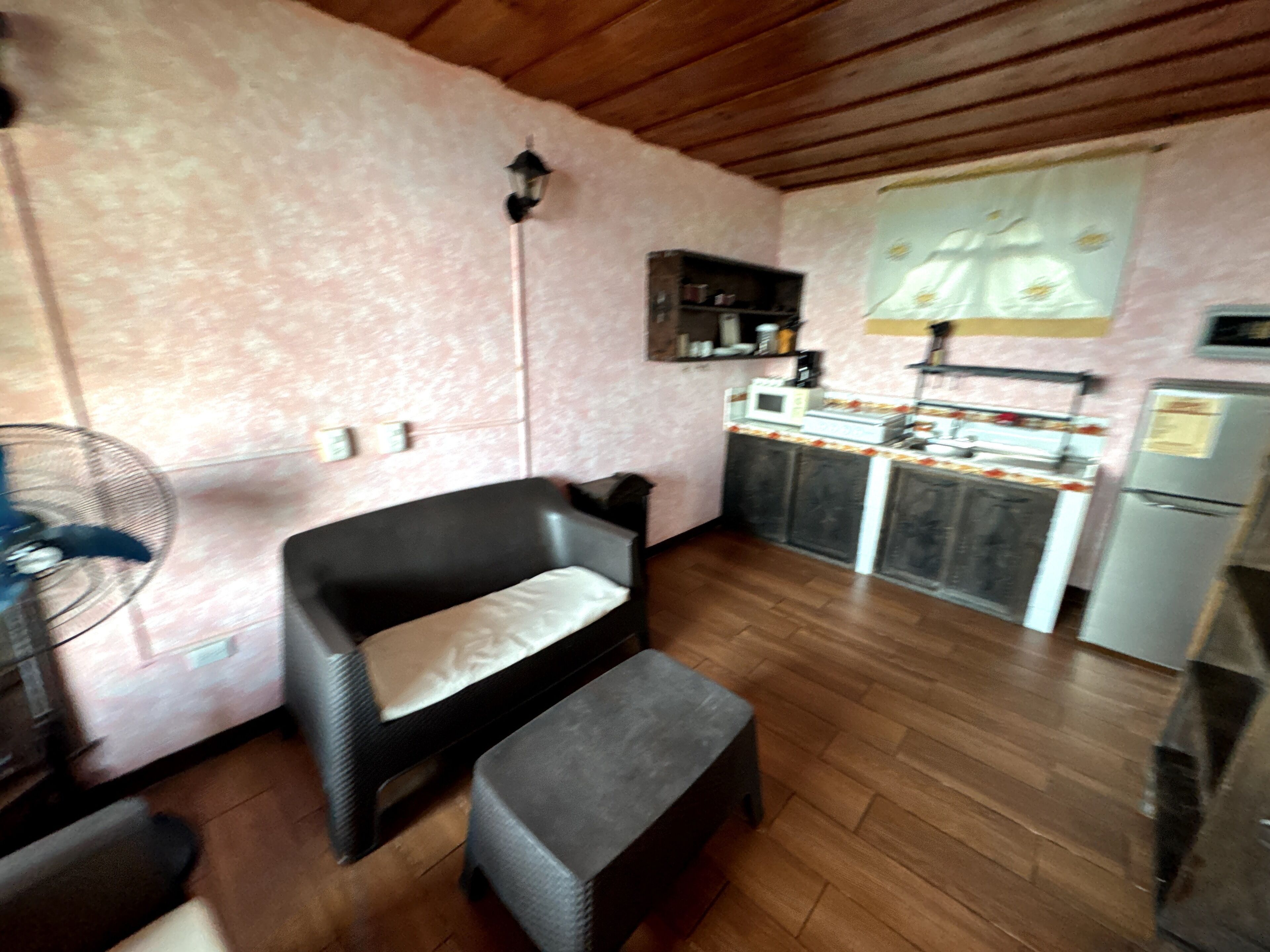 Una habitación con techo de madera, una kitchenette con microondas, estufa y refrigerador, una silla, un banco y un ventilador.