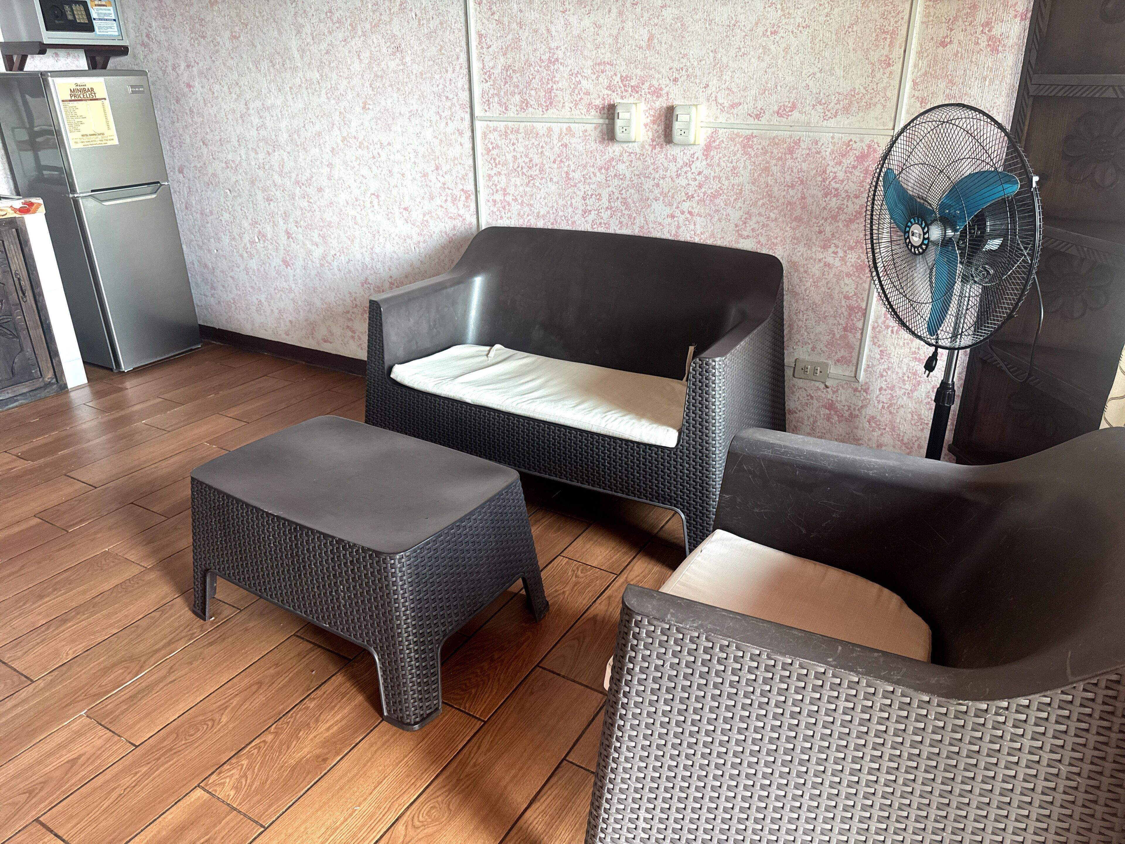 A room with a sofa, a matching ottoman, a standing fan, and a refrigerator.
