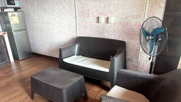 A room with a sofa, a matching ottoman, a standing fan, and a refrigerator.