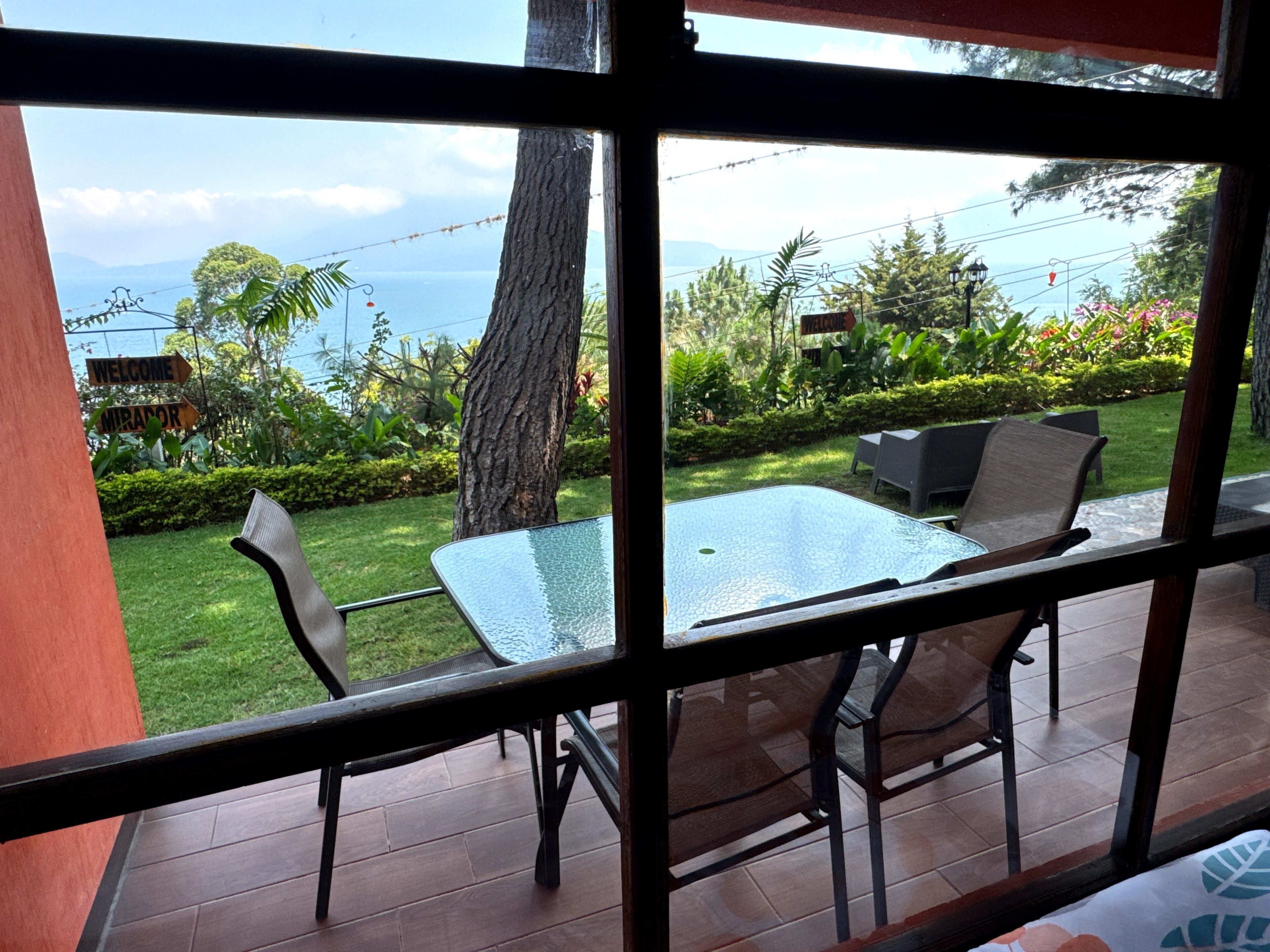 Une terrasse avec une table et des chaises en verre, une vue sur un jardin luxuriant et une pancarte sur laquelle on peut lire « Bienvenue ».