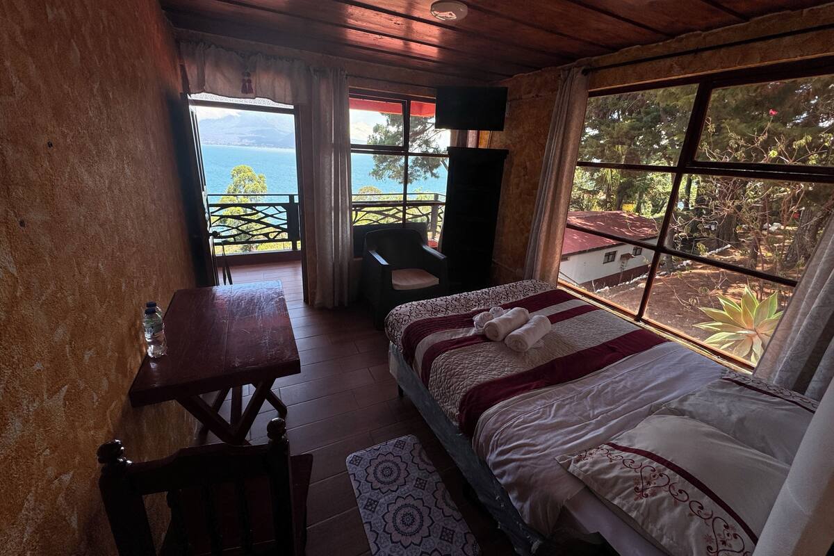 A bedroom with a bed, a desk, a chair, and a balcony with a view of the sea.