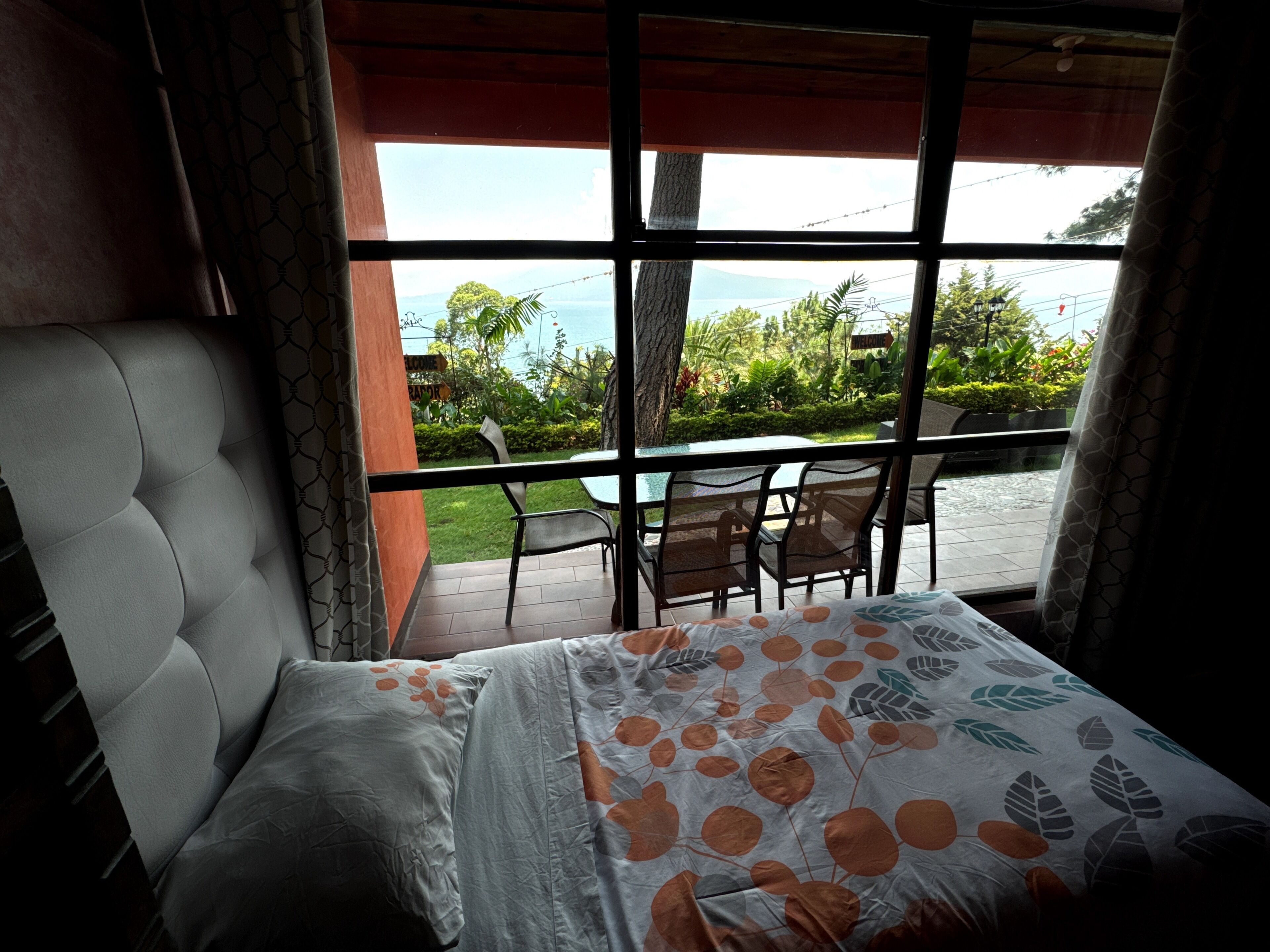 Una habitación con cama, ventana que da a una piscina y follaje, y balcón con sillas y una mesa.