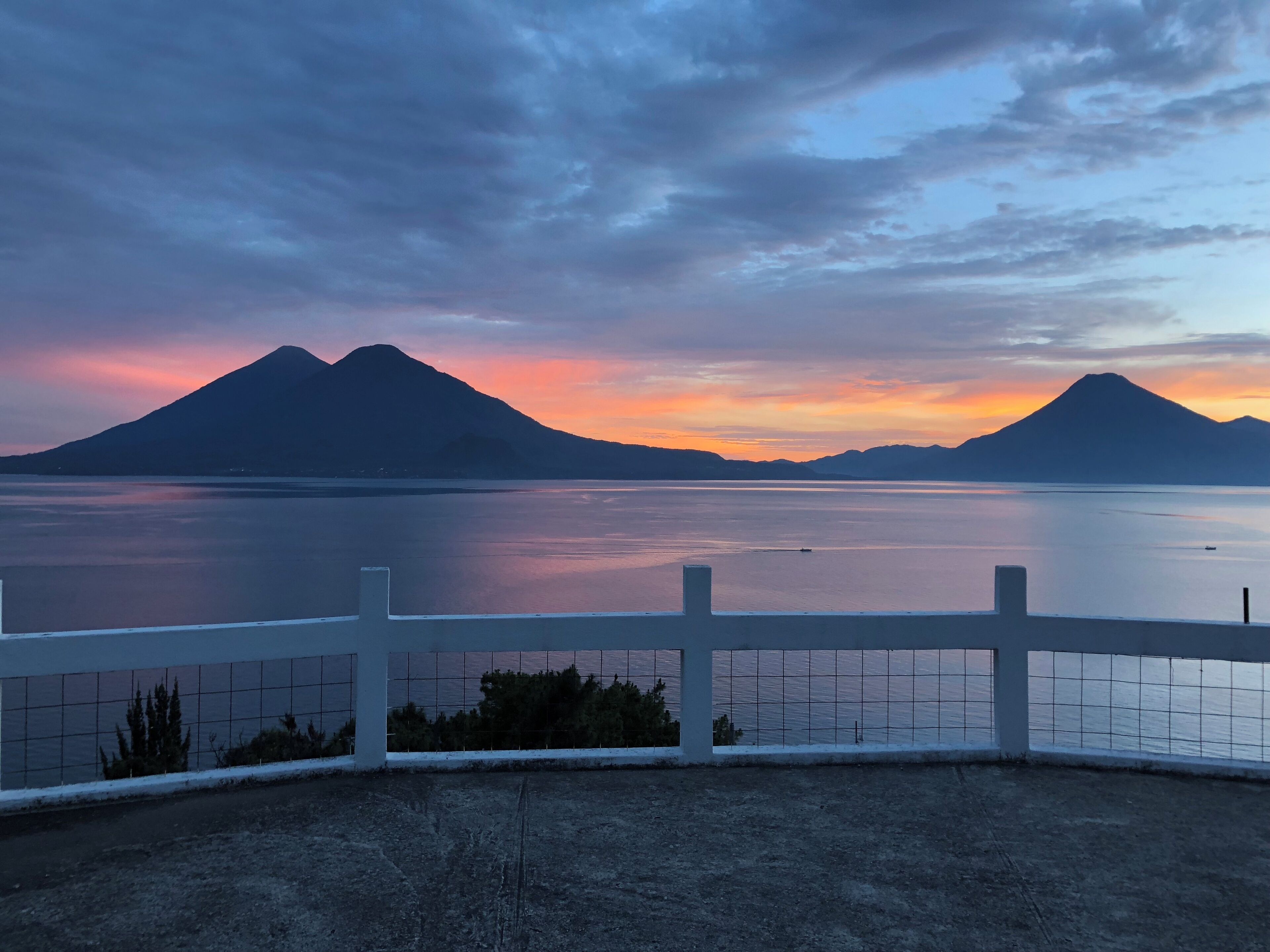 Vista de un lago tranquilo con montañas al fondo y una valla blanca en primer plano.