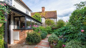 A garden pathway leading to a conservatory with a brick wall and glass doors, surrounded by lush greenery and flowering plants. - Tegfan – Fairytale Cottage on Dedham High Street (Colchester)