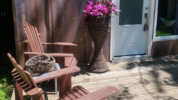 A wooden deck with a bench and chairs, a wicker planter with flowers, and a white door with a hanging wreath.