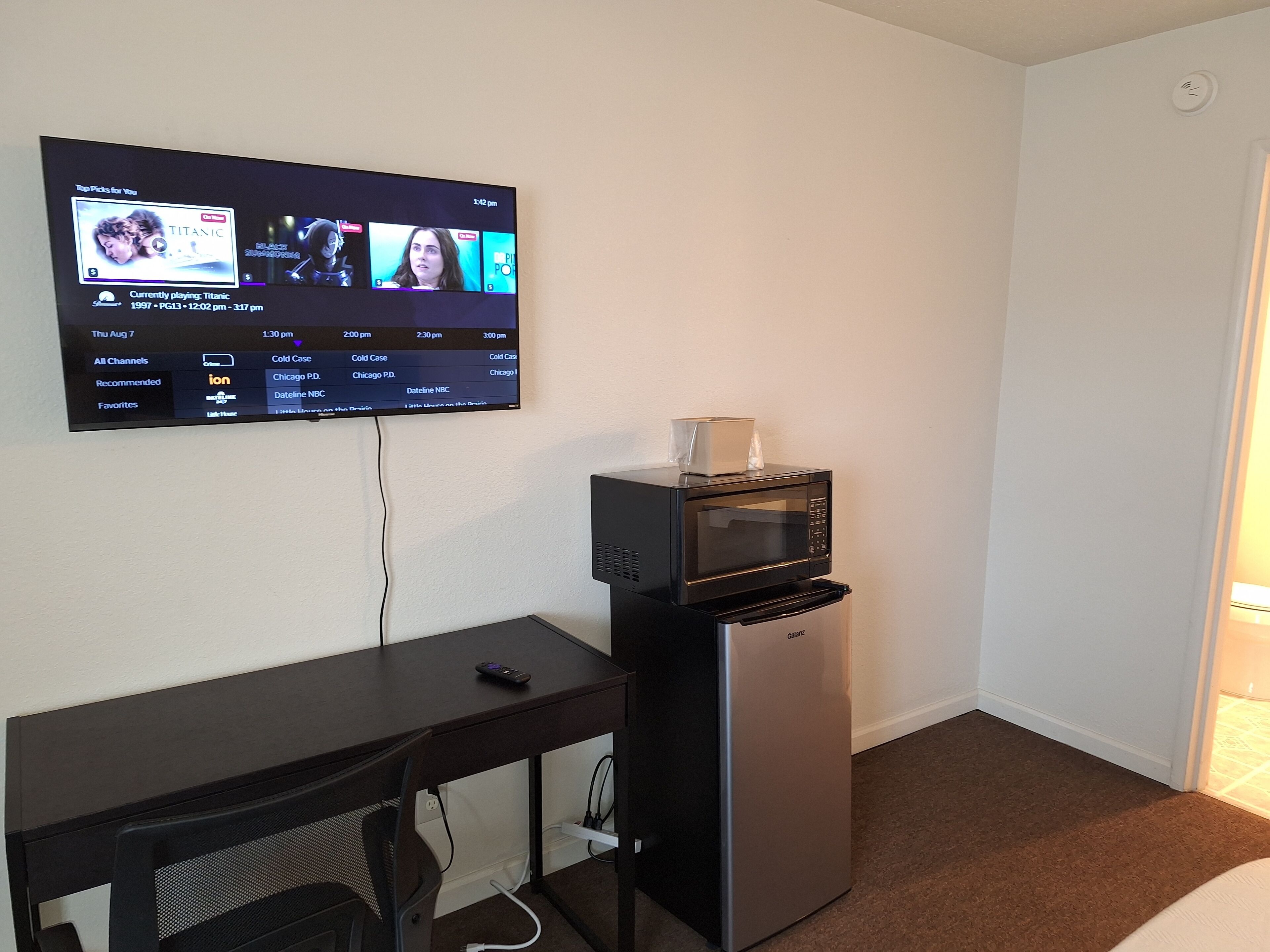 A hotel room with a flat-screen TV mounted on the wall displaying a streaming service interface, a small refrigerator, and a desk with a remote control.