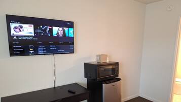 A hotel room with a flat-screen TV mounted on the wall displaying a streaming service interface, a small refrigerator, and a desk with a remote control.