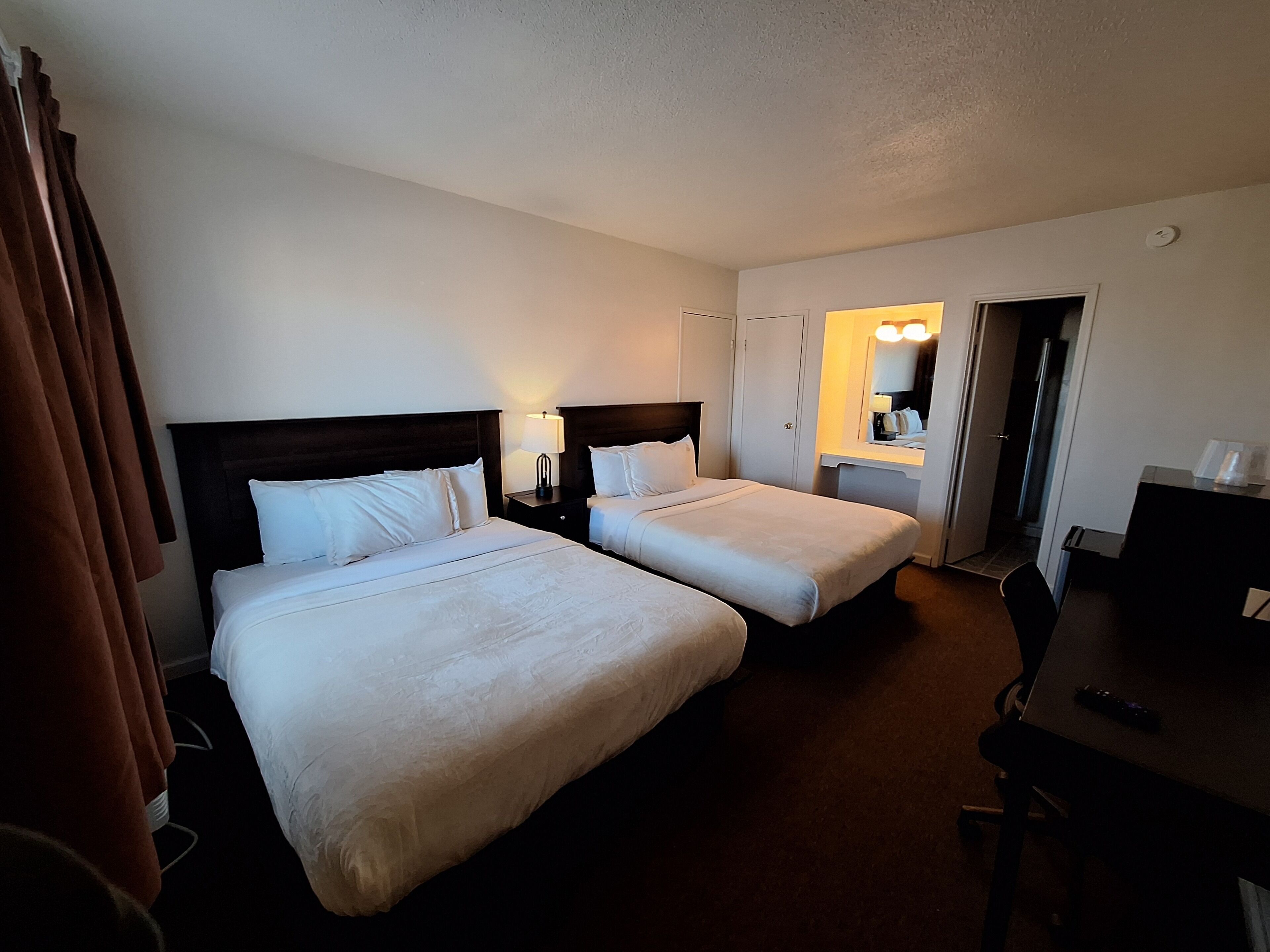 A hotel room with two beds, a desk, a chair, a wardrobe, and a bathroom visible through an open door.