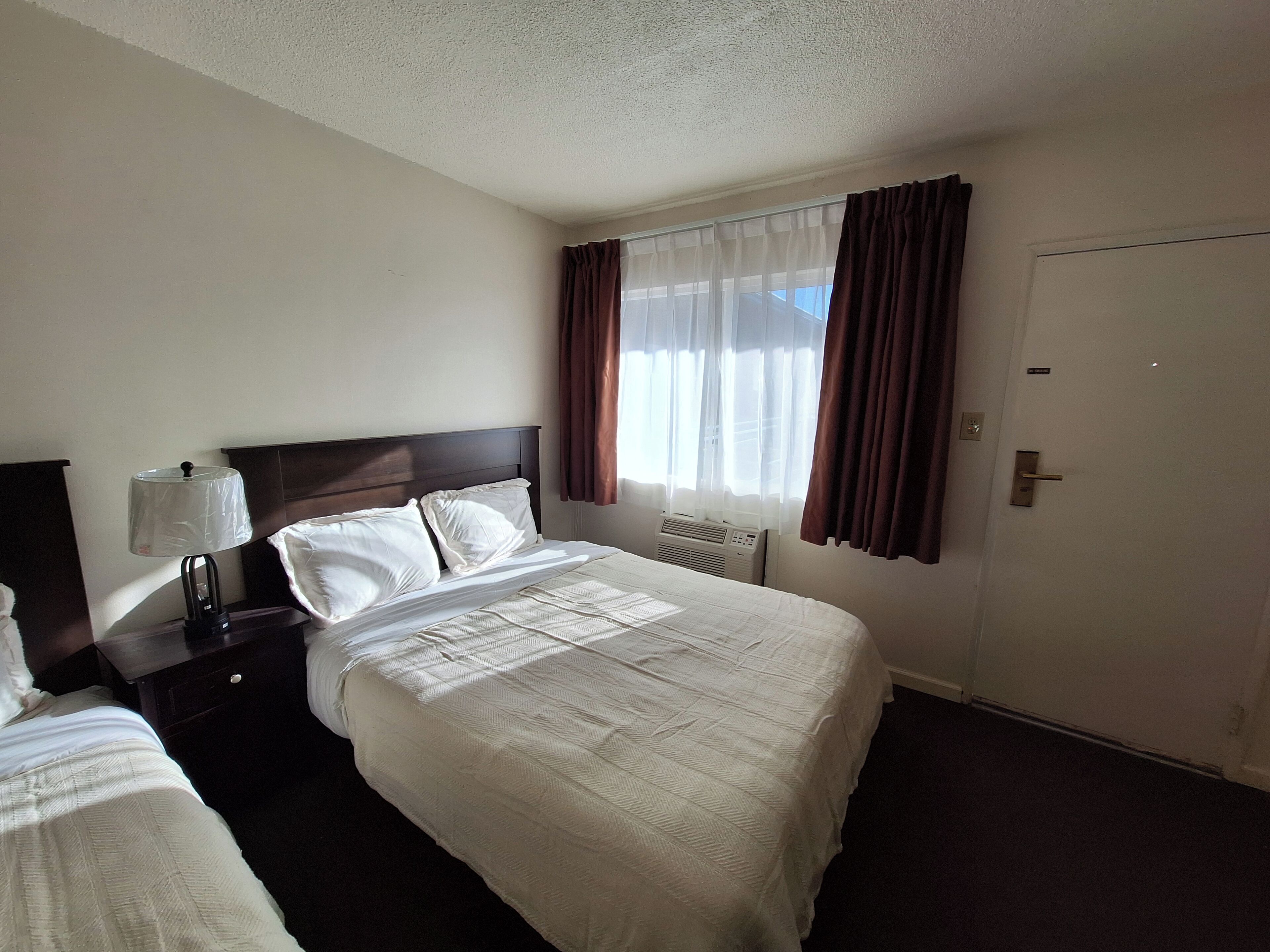 Habitación de hotel con cama, mesita de noche, lámpara, ventana con cortinas y puerta.