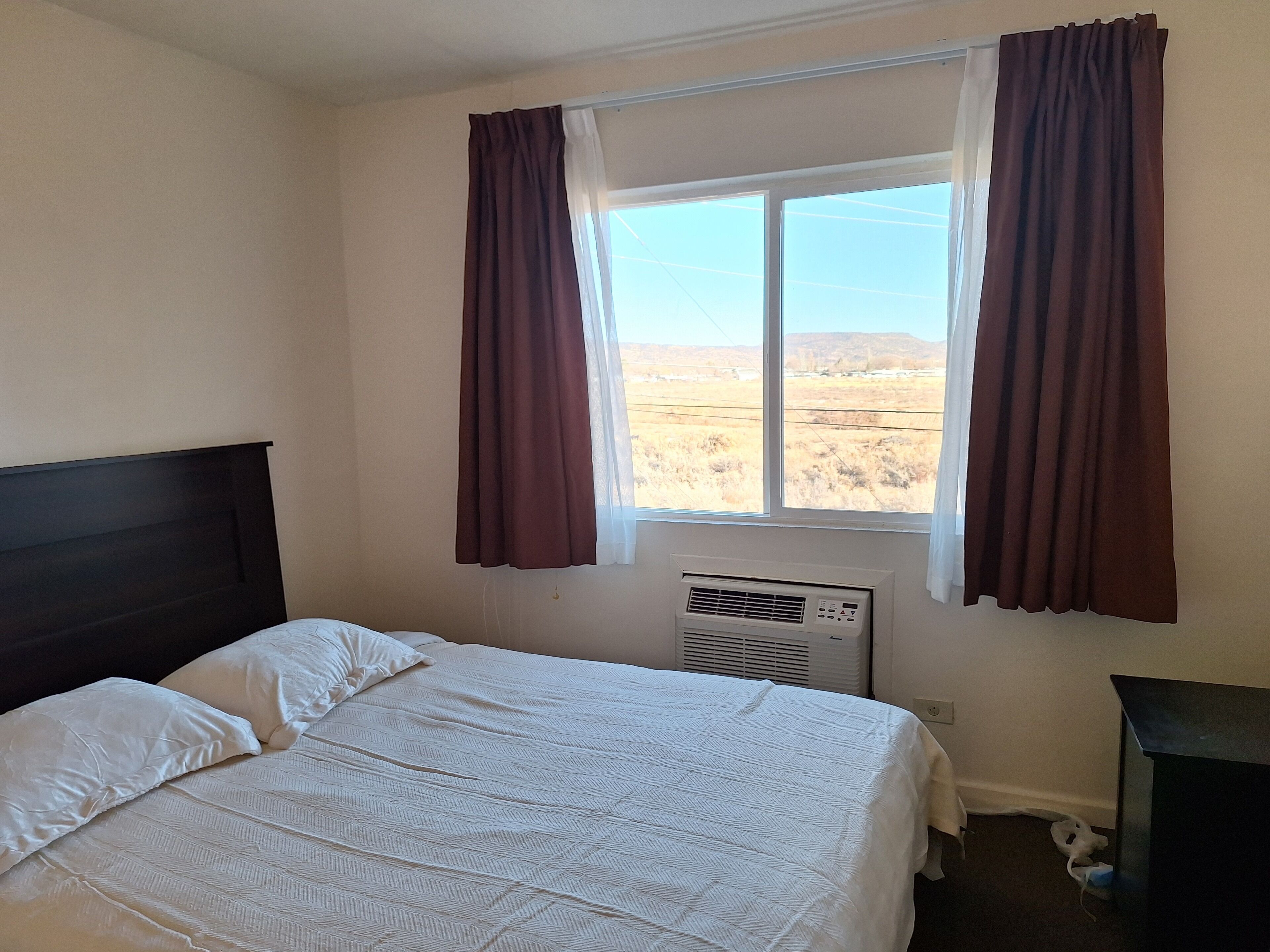 A bedroom with a bed, a window with curtains, an air conditioning unit, and a nightstand.