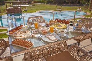 A breakfast buffet with various pastries, fruits, and beverages on a table by a pool.