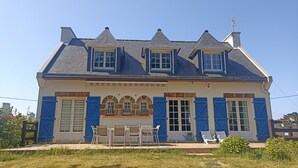 A two-story building with a gabled roof, dormer windows, and a front porch with chairs and a table. - House for 10 people in Bois du Kador – surrounded by nature (Crozon)