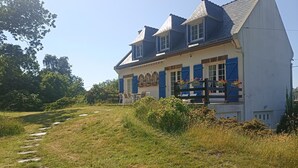 Une maison à deux étages, dotée d’un toit en pente, d’un porche à l’avant avec des chaises, et d’un chemin menant à l’entrée.