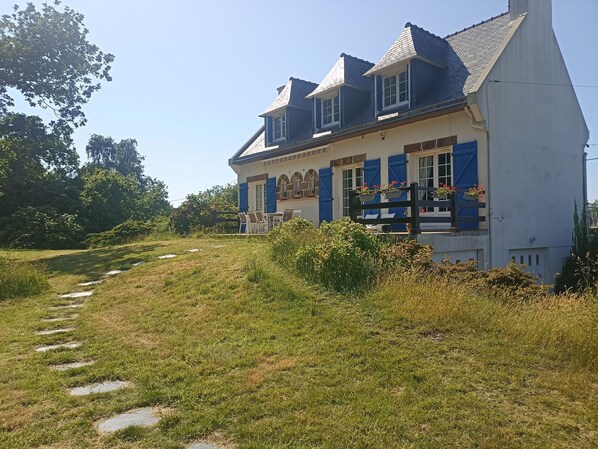 A two-story house with a sloped roof, a front porch with chairs, and a pathway leading to the entrance.