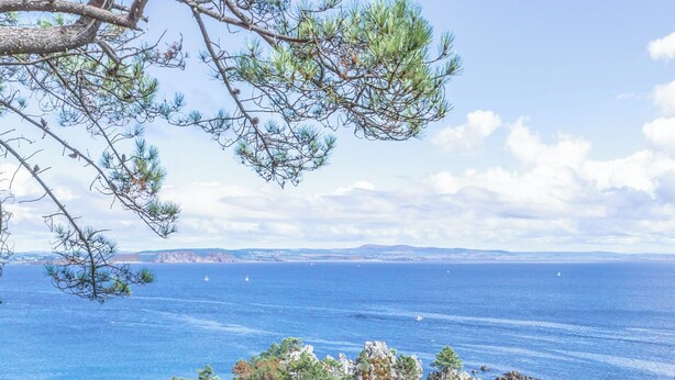 A woman sitting on a rocky ledge overlooking a beach with clear turquoise water and a rocky outcrop. - House for 10 people in Bois du Kador – surrounded by nature (Crozon)