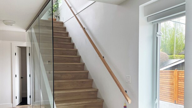 Un intérieur moderne avec un escalier en bois, des rampes de protection en verre et une grande fenêtre donnant sur l’extérieur.