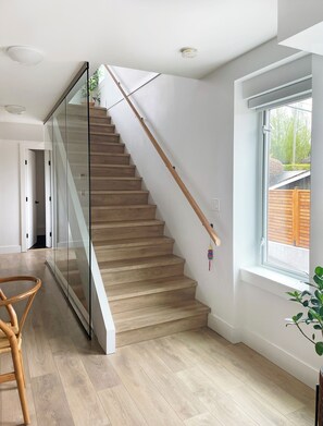Un intérieur moderne avec un escalier en bois, des rampes de protection en verre et une grande fenêtre donnant sur l’extérieur.