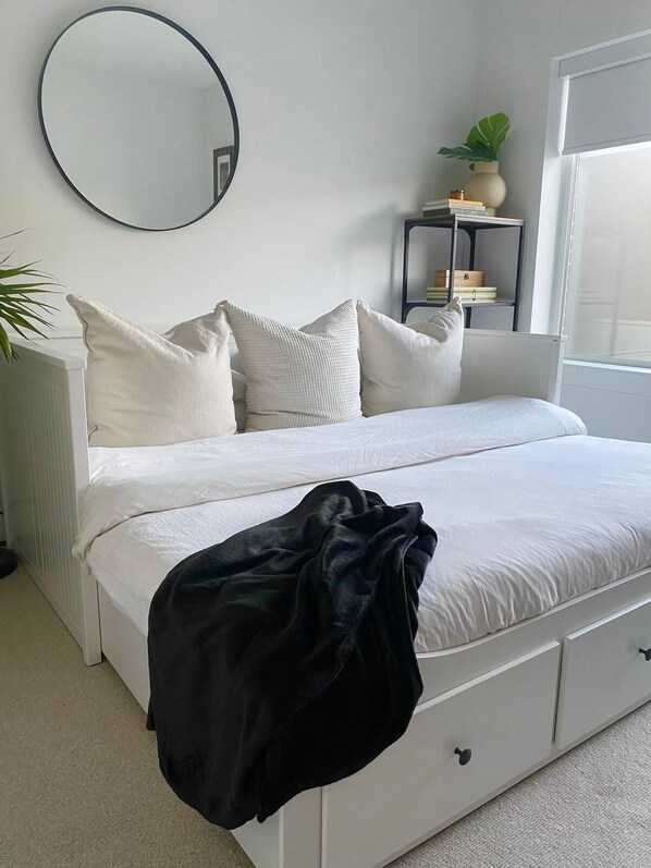Une chambre à coucher avec un lit, du linge de lit blanc, un plaid noir, un miroir rond, une plante sur la table de chevet et une petite étagère avec des livres et un vase.