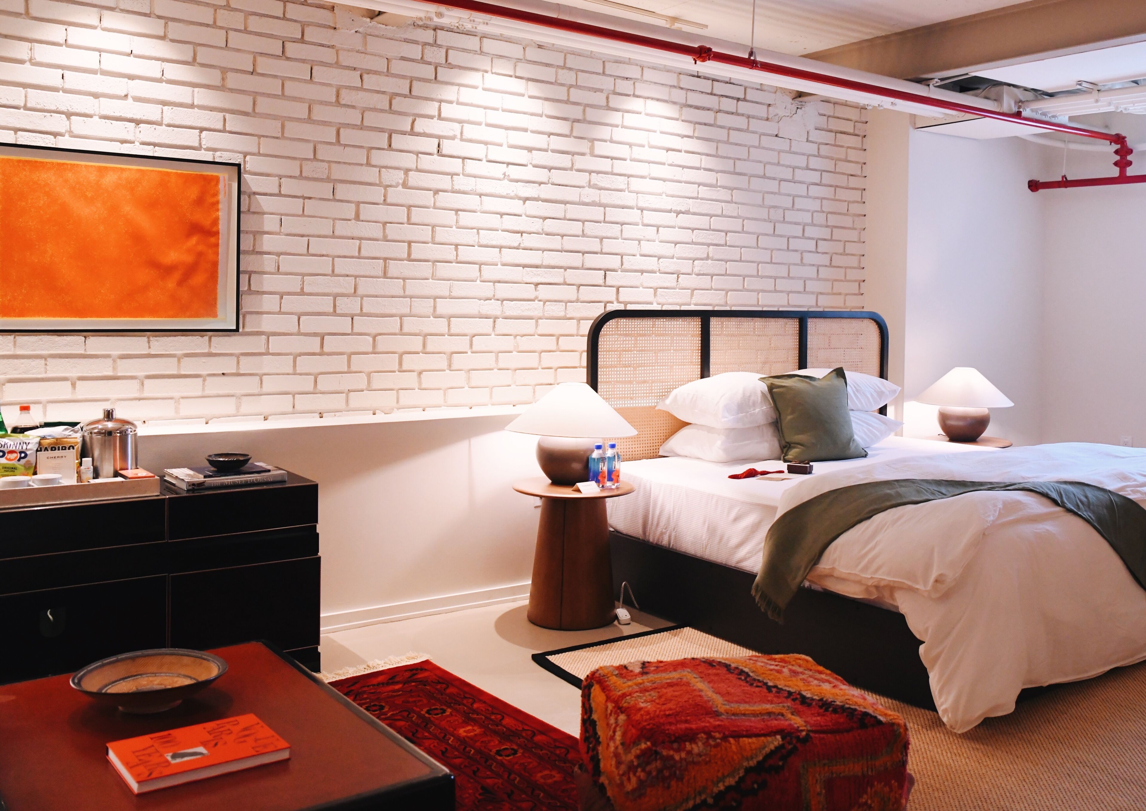 Ein modernes Hotelzimmer mit Backsteinwand, einem Bett mit weißer Bettwäsche, zwei Nachttischlampen, einem roten Teppich und einem kleinen Tisch mit einem Buch und einer Schüssel.