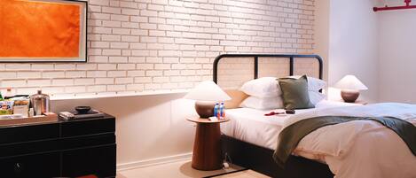 Ein modernes Hotelzimmer mit Backsteinwand, einem Bett mit weißer Bettwäsche, zwei Nachttischlampen, einem roten Teppich und einem kleinen Tisch mit einem Buch und einer Schüssel.