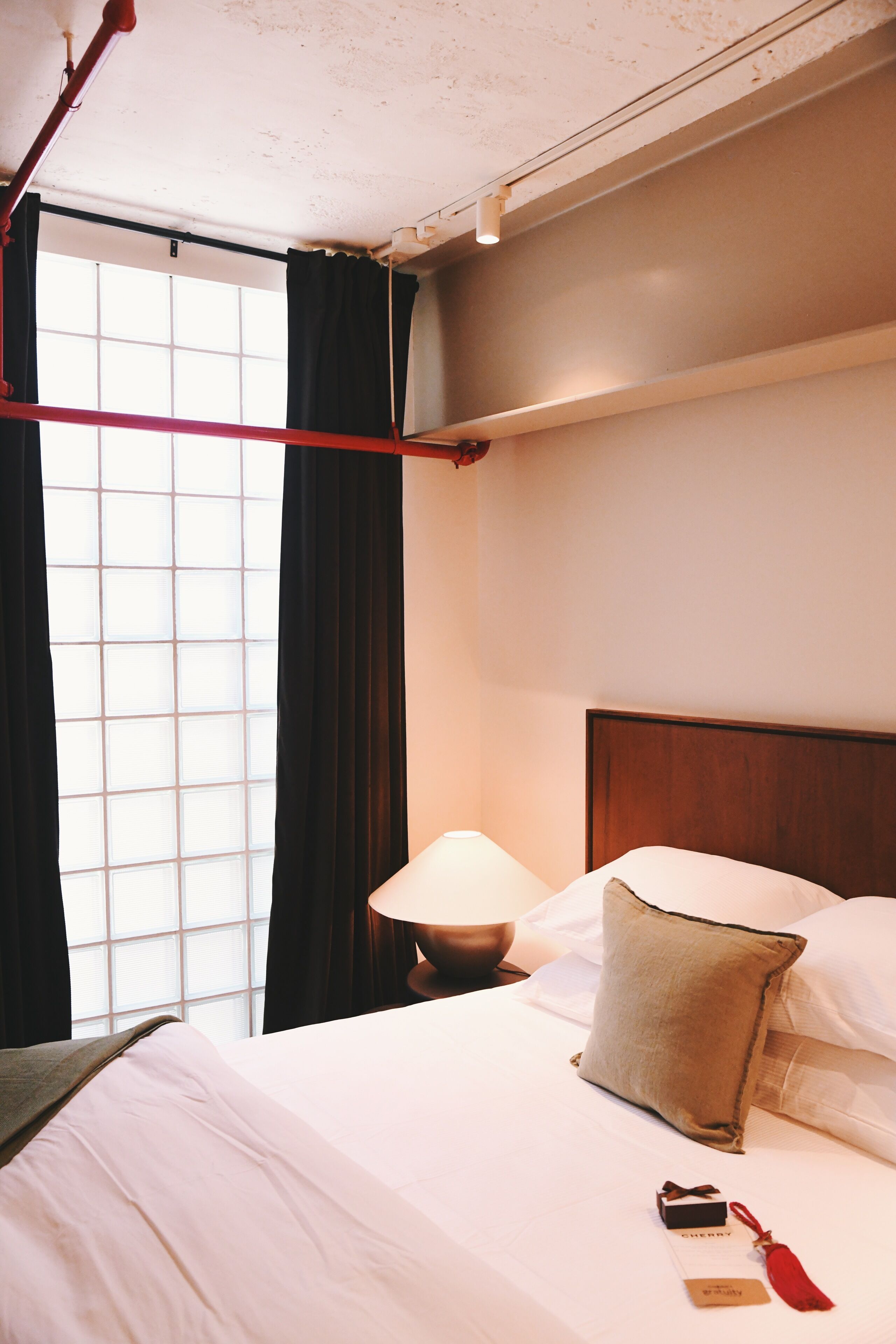 Una habitación de hotel con cama, mesita de noche, lámpara y ventana con cortinas.