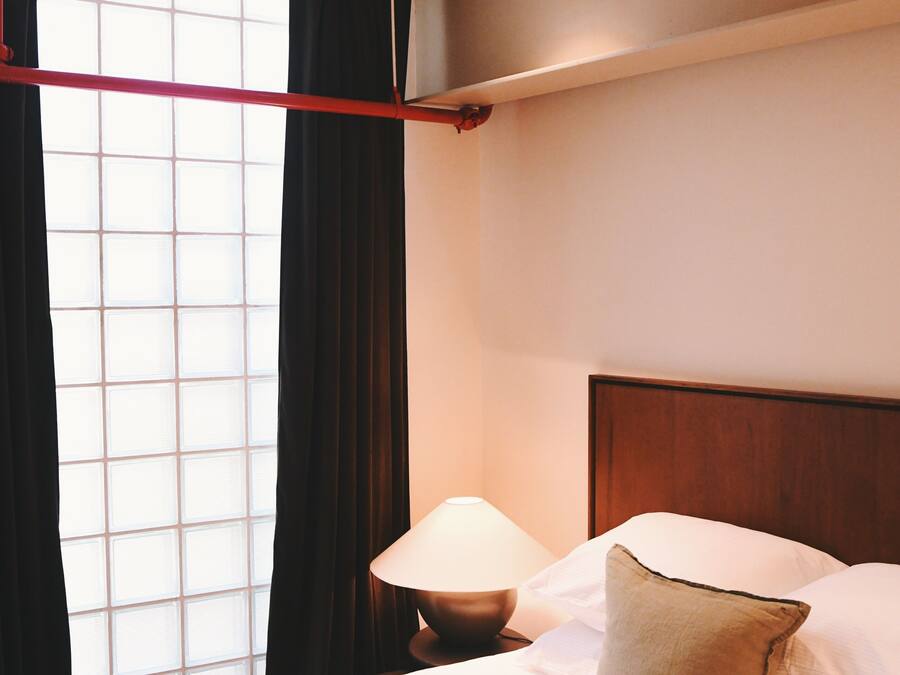 Una habitación de hotel con cama, mesita de noche, lámpara y ventana con cortinas.