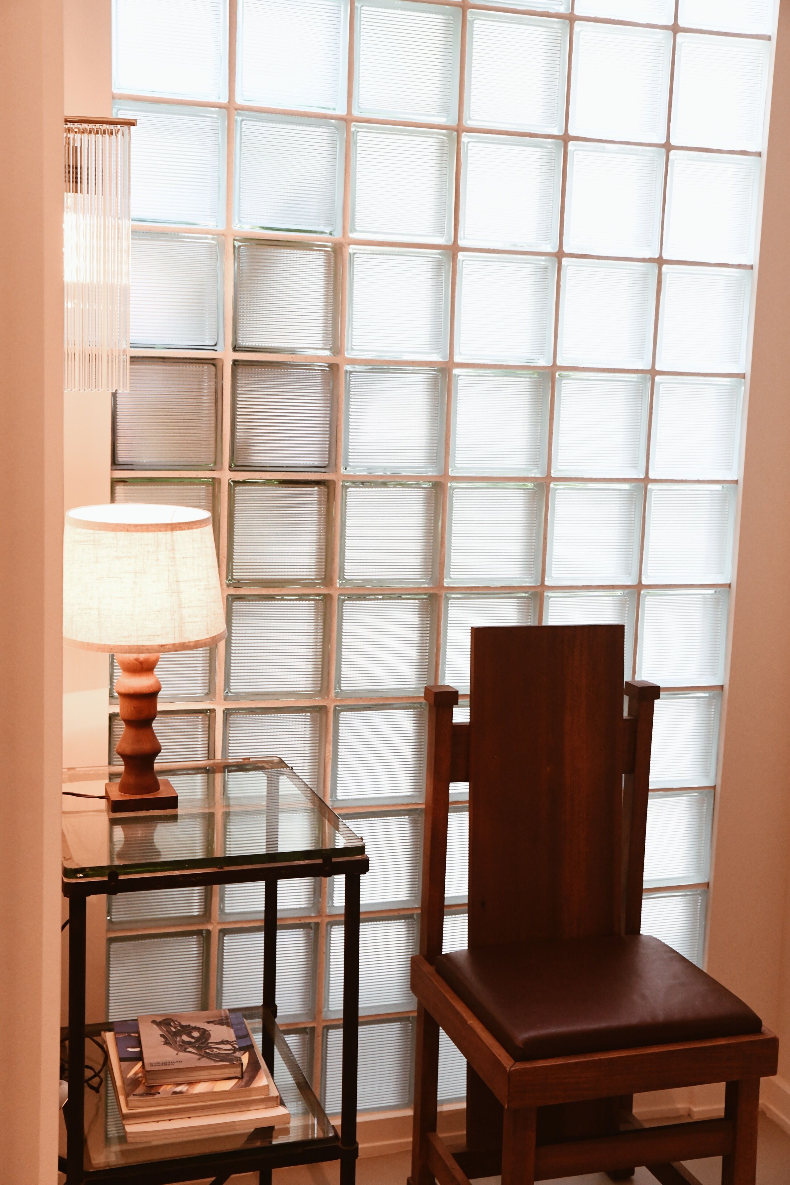 Une pièce avec une table en verre sur laquelle se trouvent une lampe et des livres, une chaise en bois et une fenêtre avec des stores.