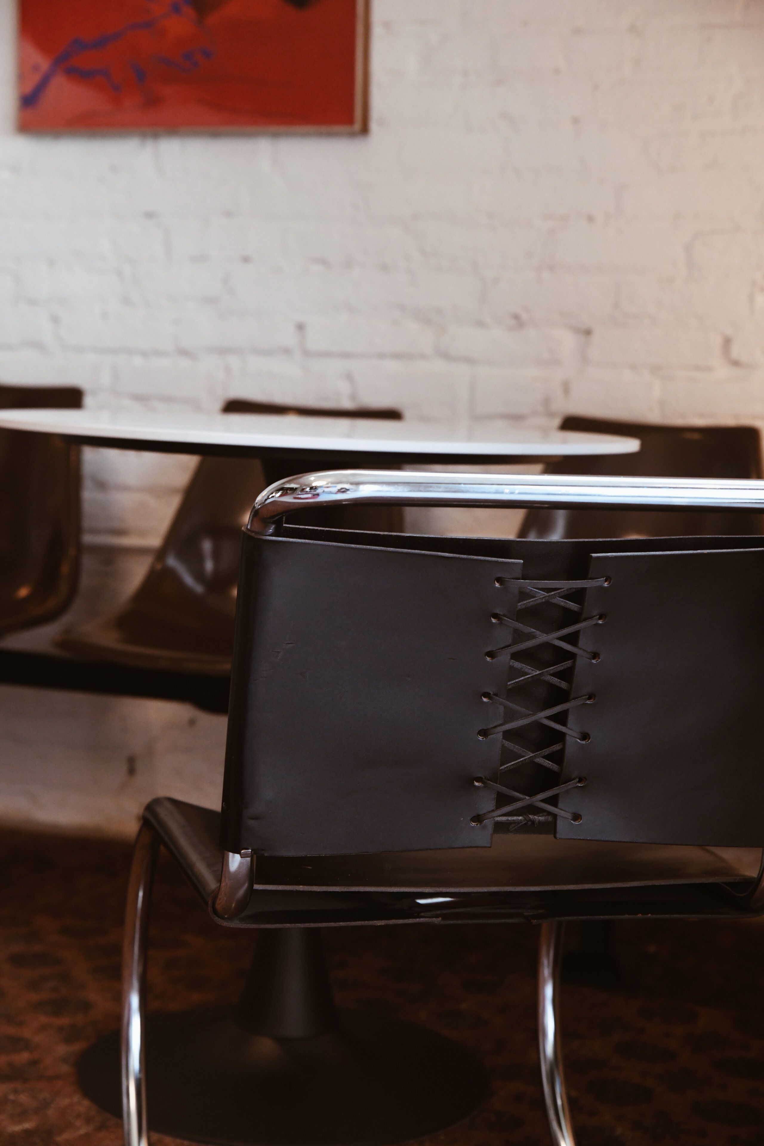A black chair with a metal frame and a laced backrest, positioned in front of a white brick wall and a table with a reflective surface.