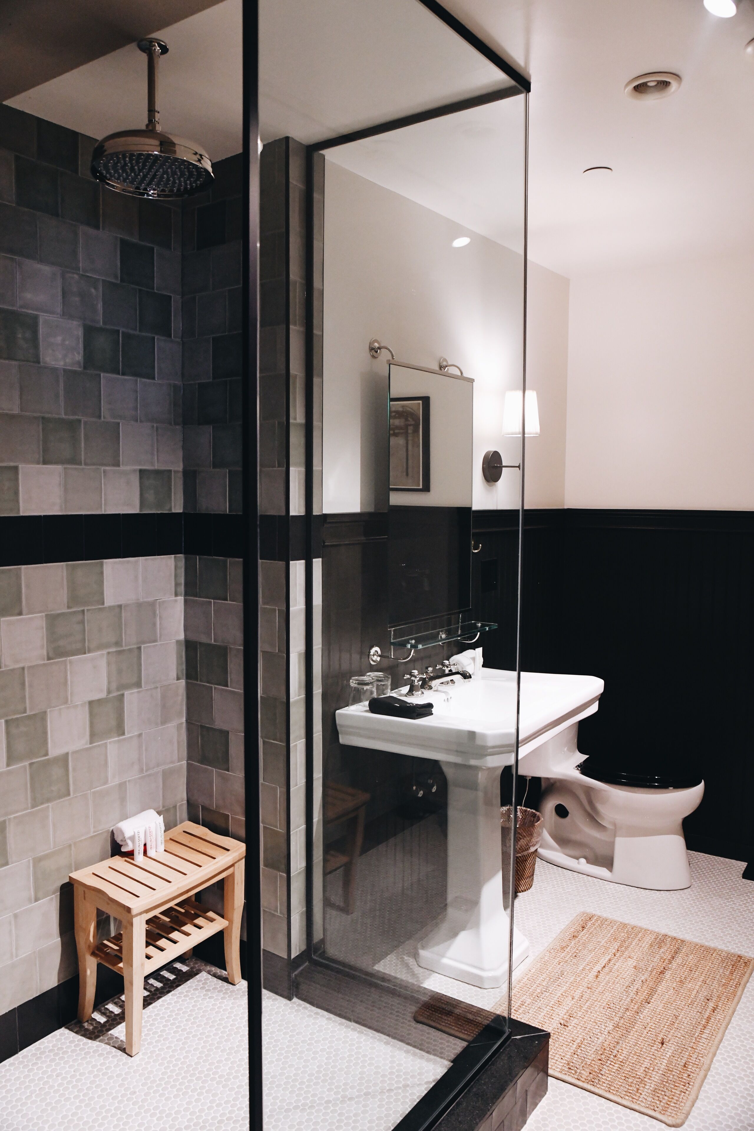 A modern bathroom with a glass shower enclosure, a wooden stool, a white sink, and a black bathtub.