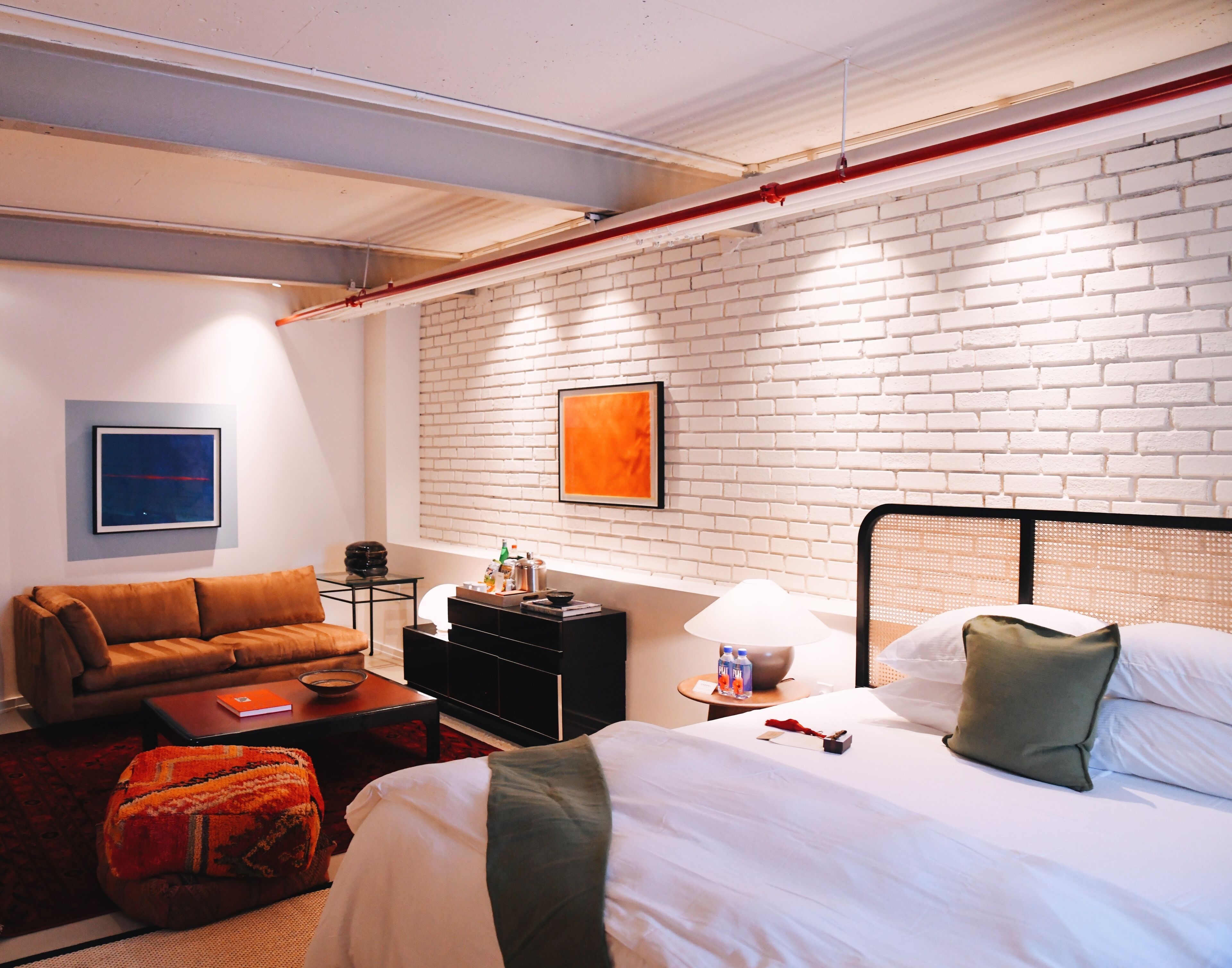 Una habitación de hotel con una pared de ladrillo blanco, una cama con almohadas verdes, un sofá marrón, una mesita con una lámpara y un cuadro en la pared.