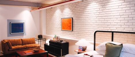 Una habitación de hotel con una pared de ladrillo blanco, una cama con almohadas verdes, un sofá marrón, una mesita con una lámpara y un cuadro en la pared.