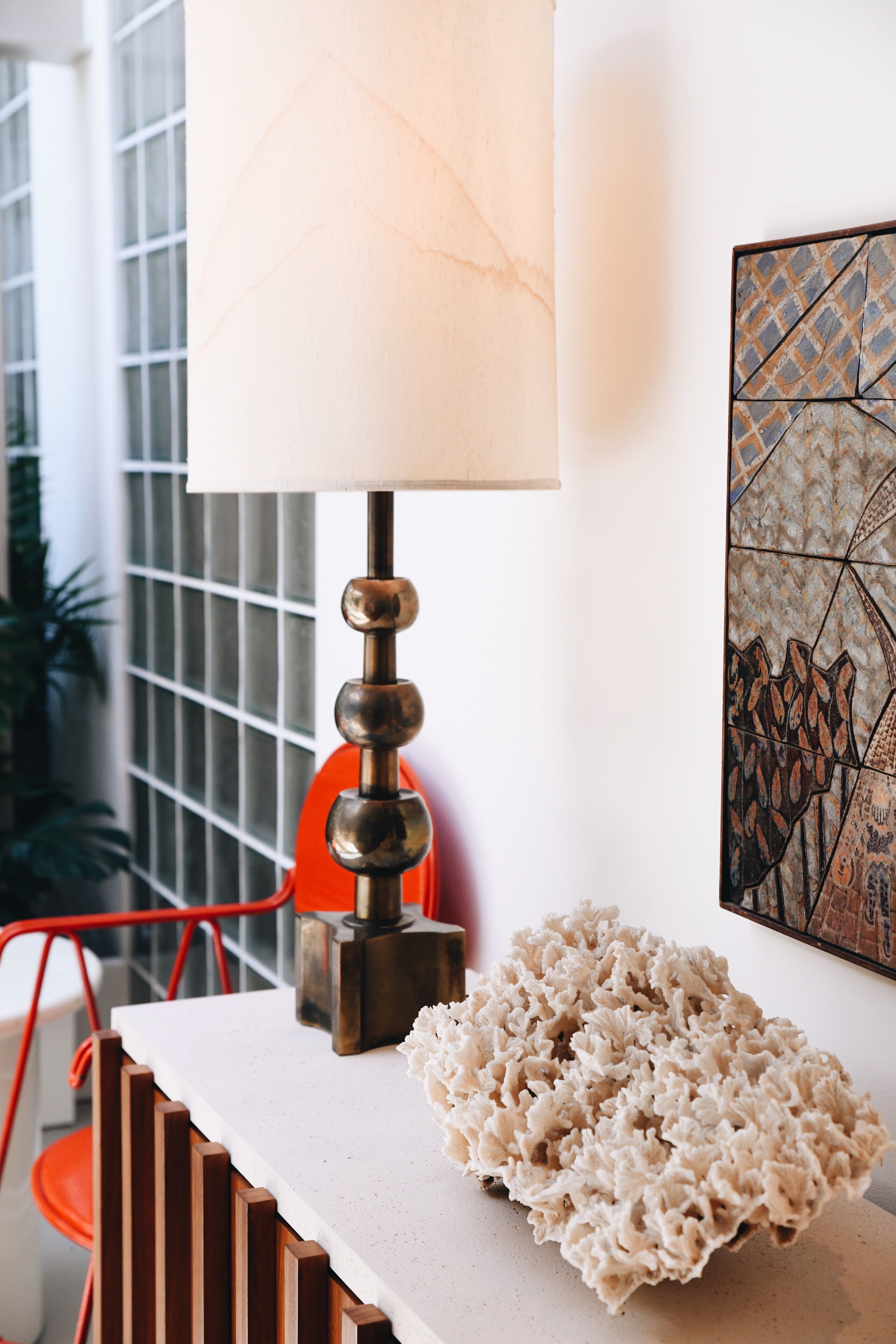 Ein modernes Interieur mit einem weißen Tisch, einem dekorativen Korallenstück, einer Lampe und einem geometrischen Kunstwerk an der Wand.