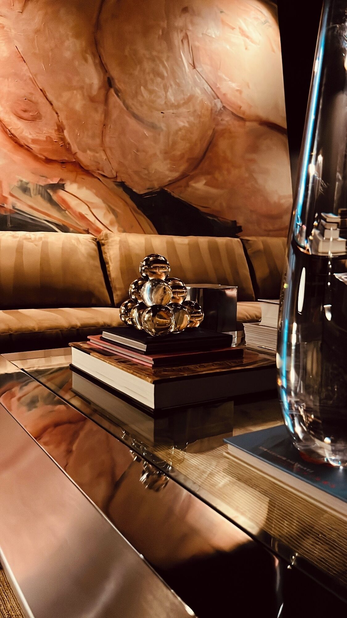 A modern living room with a glass coffee table, a stack of books, decorative glass spheres, and a large abstract painting on the wall.