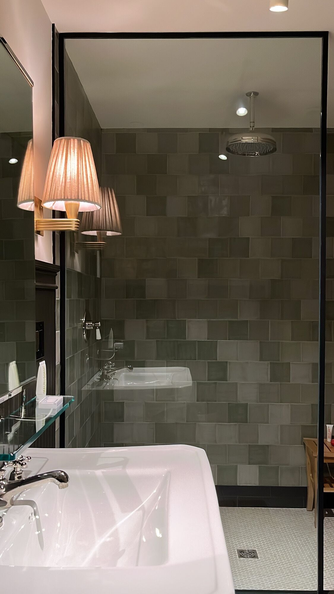 A modern bathroom with a large mirror, two wall-mounted lamps, a glass shower enclosure, and a white sink.