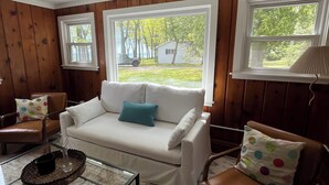 A living room with a white sofa, a brown leather chair, a glass coffee table, and a view of a house through large windows. - Charming Lake Huron cottage (Tawas City)