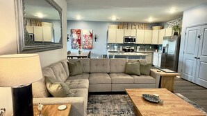 A modern living room with a sectional sofa, a coffee table, a lamp, and a rug. There is a kitchen in the background with cabinets and appliances. - NEW Putt Chip&Chill 3BRTownhouse NMB SportsComplex (little river)