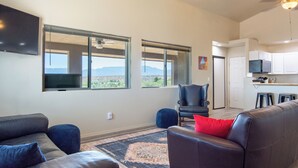 A spacious living room with a large flat-screen TV, a leather sofa, a blue armchair, and a red pillow. There are two large windows offering a view of the mountains. - Private Wine Country Retreat • 2 Suites & 2 Hot Tubs (Cornville)