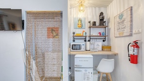 Una cocina compacta con microondas, cafetera y un refrigerador pequeño. Hay una silla blanca y un extintor de incendios montado en la pared.