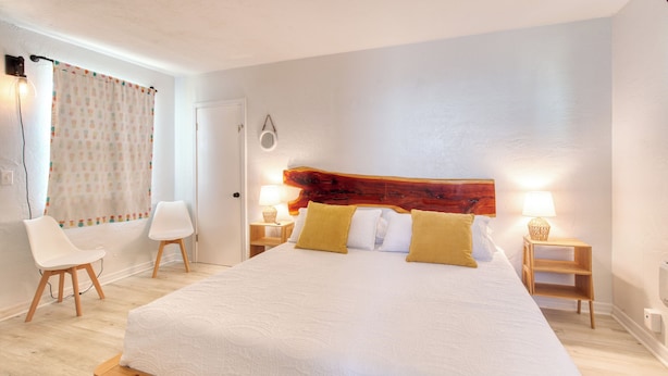 Una habitación de hotel con una cama grande, dos mesitas de noche con lámparas, un cabecero de madera, una puerta blanca y dos sillas blancas.