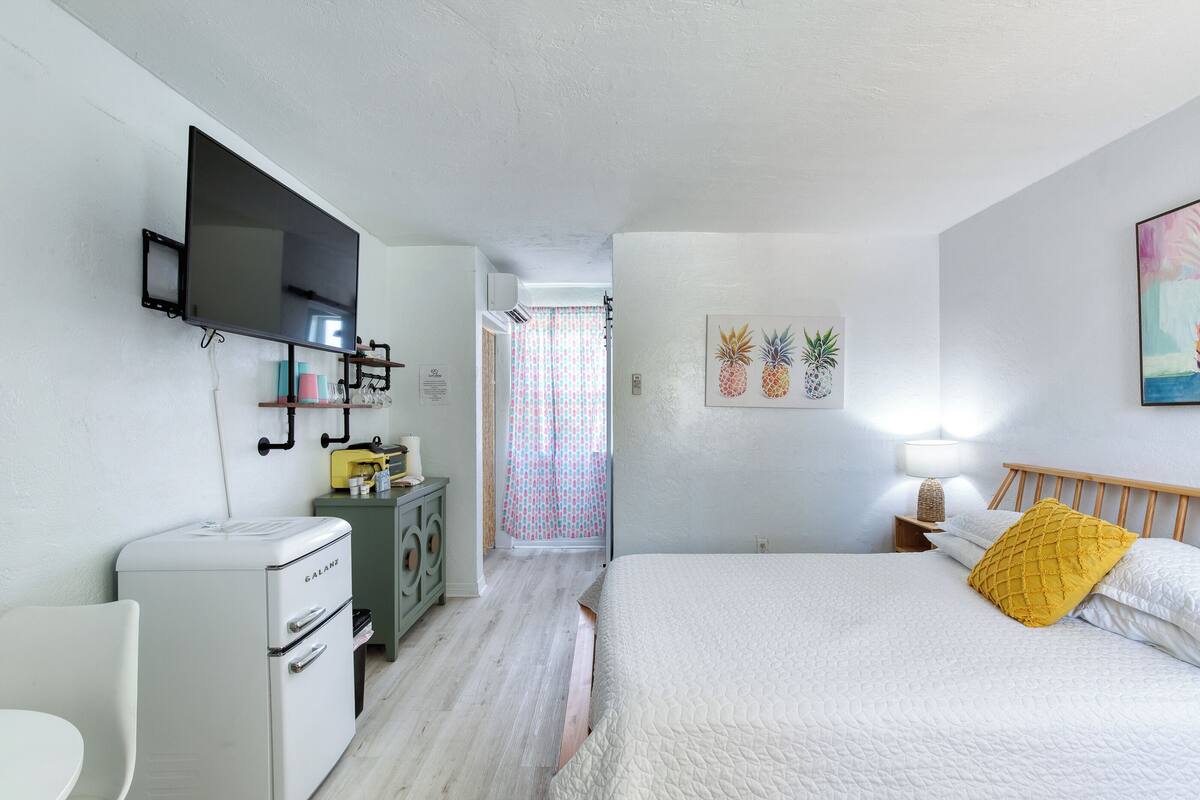 A bedroom with a bed, a TV mounted on the wall, a small kitchenette, and a bathroom visible through a door.