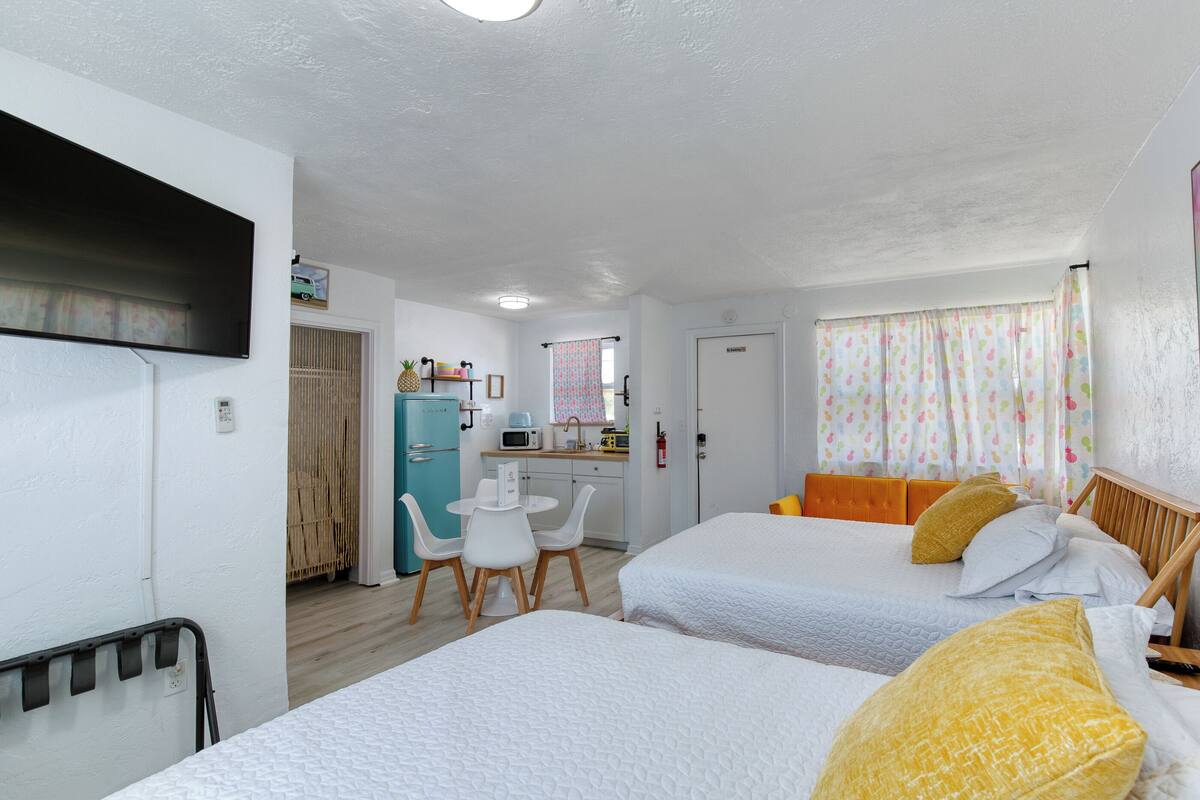 A room with two beds, a small kitchenette, a dining area, and a bathroom.