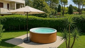 Un jacuzzi en un jardín con sombrilla de patio, rodeado de setos cuidados y una palmera.