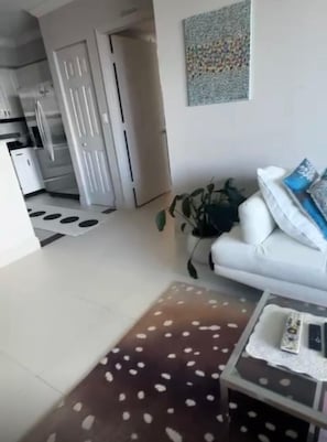 Un salon avec un canapé blanc, une table basse, un tapis à motifs, une plante et, au fond, une cuisine.