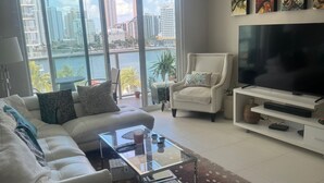 A modern living room with a white sofa, a glass coffee table, and a flat-screen TV on a white stand. Large windows offer a view of a city skyline and a body of water. - Modern 2BR Apartment in Brickell, Central w Sea View (Miami)