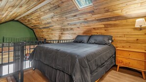 A bedroom with a wooden ceiling, a bed with a grey bedspread, a nightstand, and a window with a view of trees.