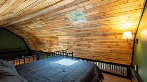 Ein Zimmer mit geneigter Holzwand, einem Bett mit dunkler Bettdecke, einem kleinen Fenster und einer Wandlampe.